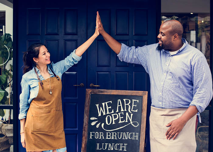 Woman and man giving high five in front of a cafe with a sign saying Open for business