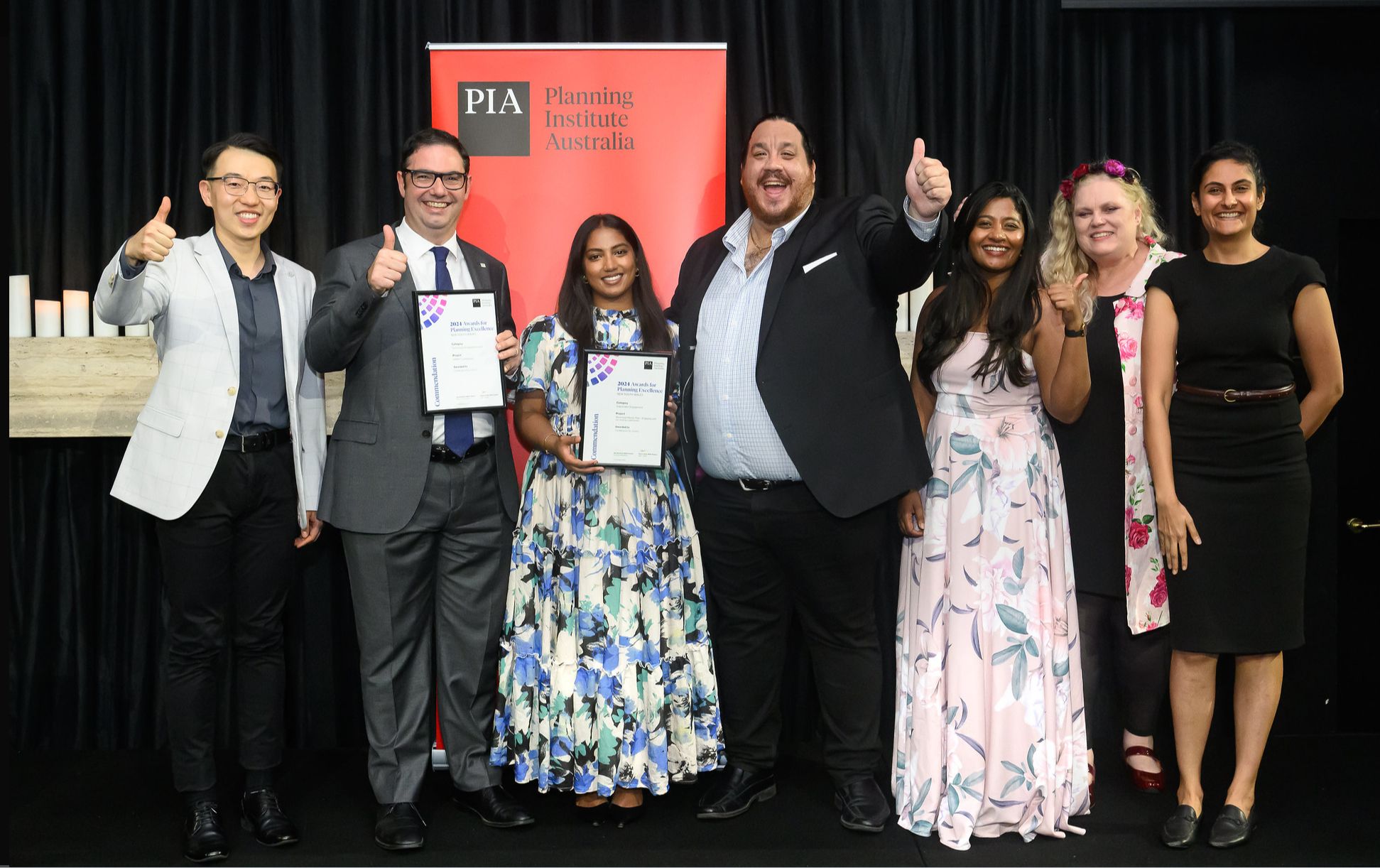 Group of 3 men and 4 women at awards ceremony holding up certificates giving thumbs up sign